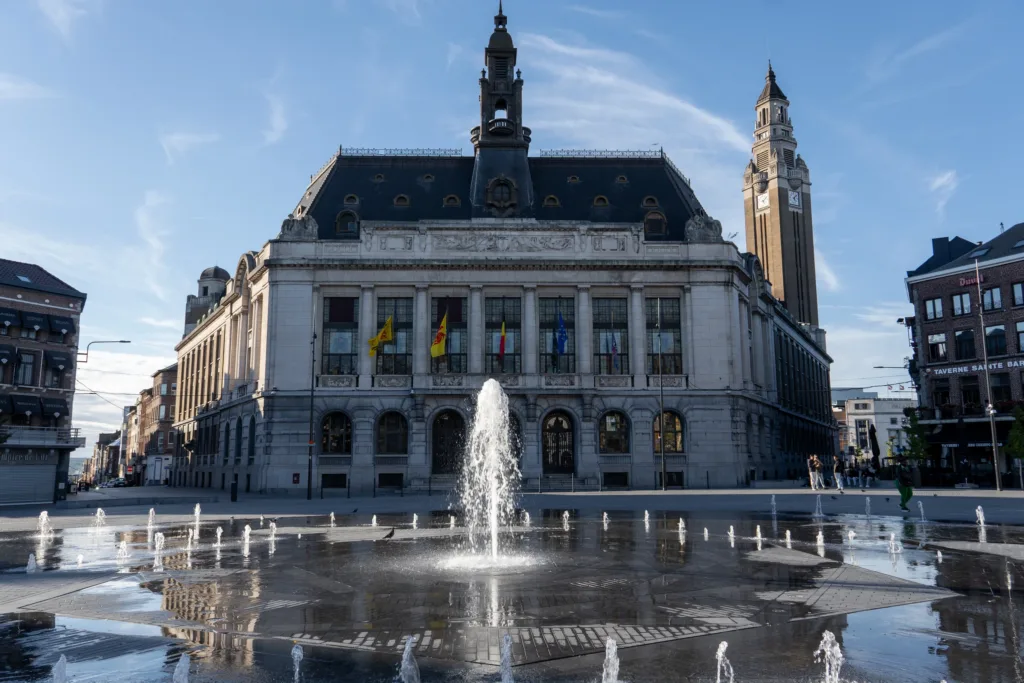 Fountain jets on Place Charles II in front of Charleroi's Town Hall and its UNESCO-listed belfry, under a clear evening sky.