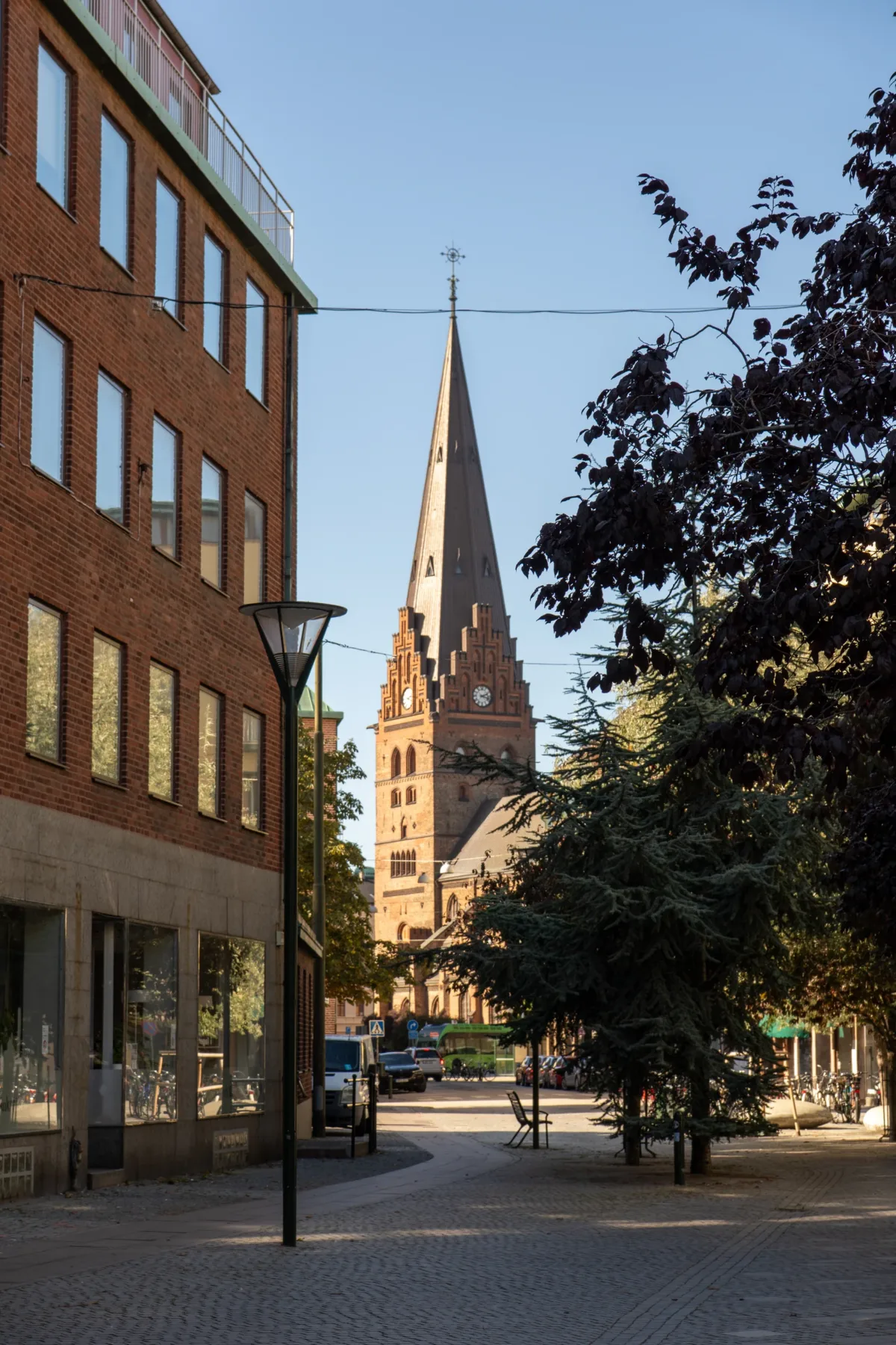 A brick church spire rises above a cobblestone street framed by trees and a modern brick building in Malmo, Sweden, under a clear blue sky.
