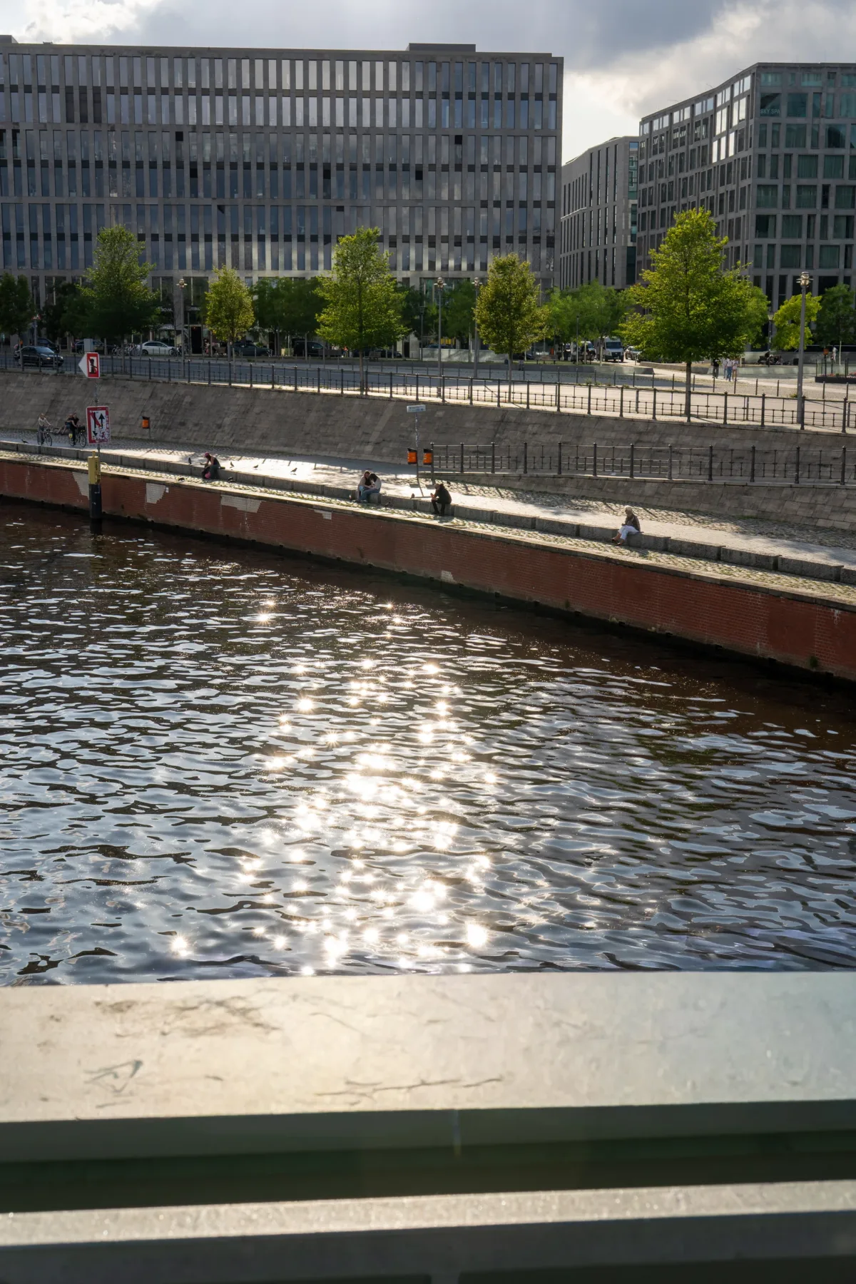 Sunlit water reflects clouds above as people sit along a riverbank beside modern glass buildings in Berlin.