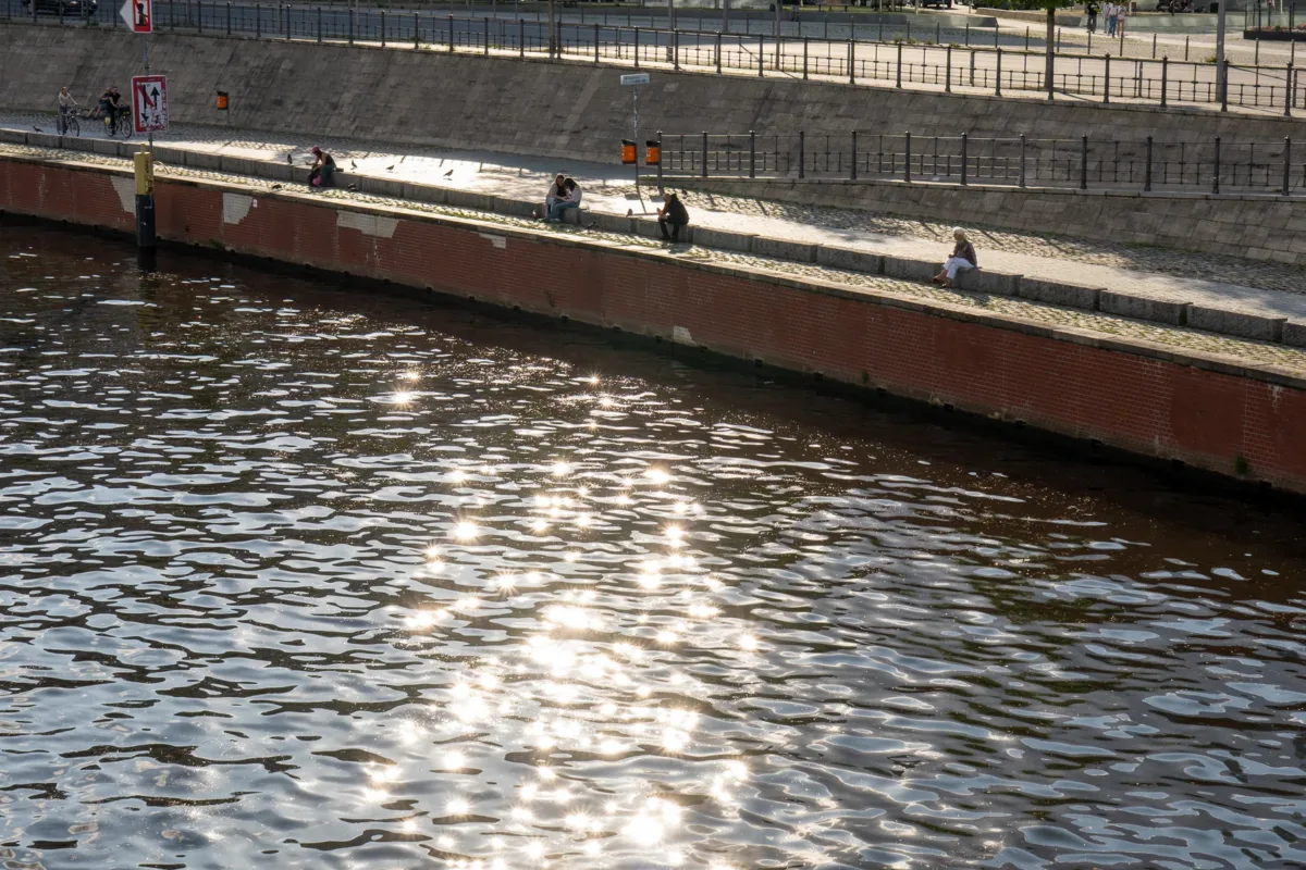 Sunlit water reflecting clouds with people sitting on riverbank beside modern glass buildings in summer midday, Berlin, Germany