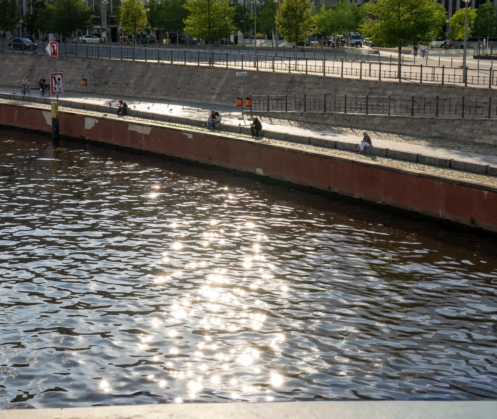 Sunlit water reflects clouds above as people sit along a riverbank beside modern glass buildings in Berlin.